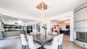 A spacious dining area with a glass-top table surrounded by white upholstered chairs, a modern kitchen in the background, and a fireplace with a stone surround. - Ayia Napa Panorama Villa (Ayia Napa)