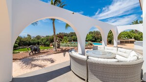 A covered outdoor seating area with wicker furniture and a view of a landscaped garden and pool. - Ayia Napa Panorama Villa (Ayia Napa)