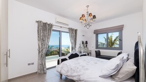 A bedroom with a large bed, a chandelier, a view of the sea, and a window with curtains. - Ayia Napa Panorama Villa (Ayia Napa)