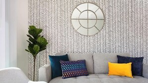 Un salon avec un papier peint à motifs en chevrons, un miroir rond et un canapé gris agrémenté de coussins colorés.