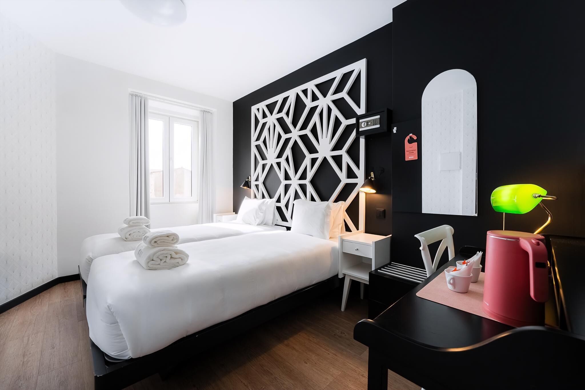 A hotel room with two beds, a desk, a chair, a lamp, and a kettle.