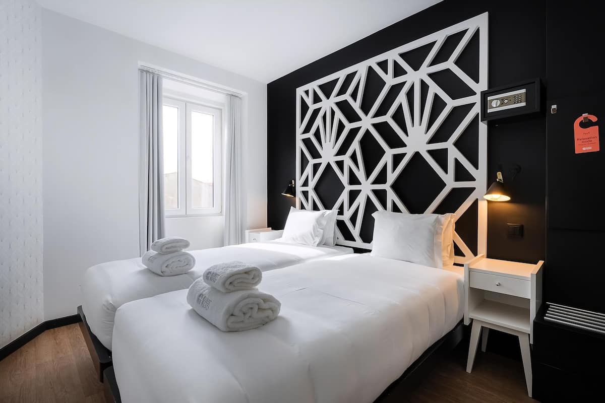 A hotel room with two beds, a geometric wall panel, a bedside table, and a window.