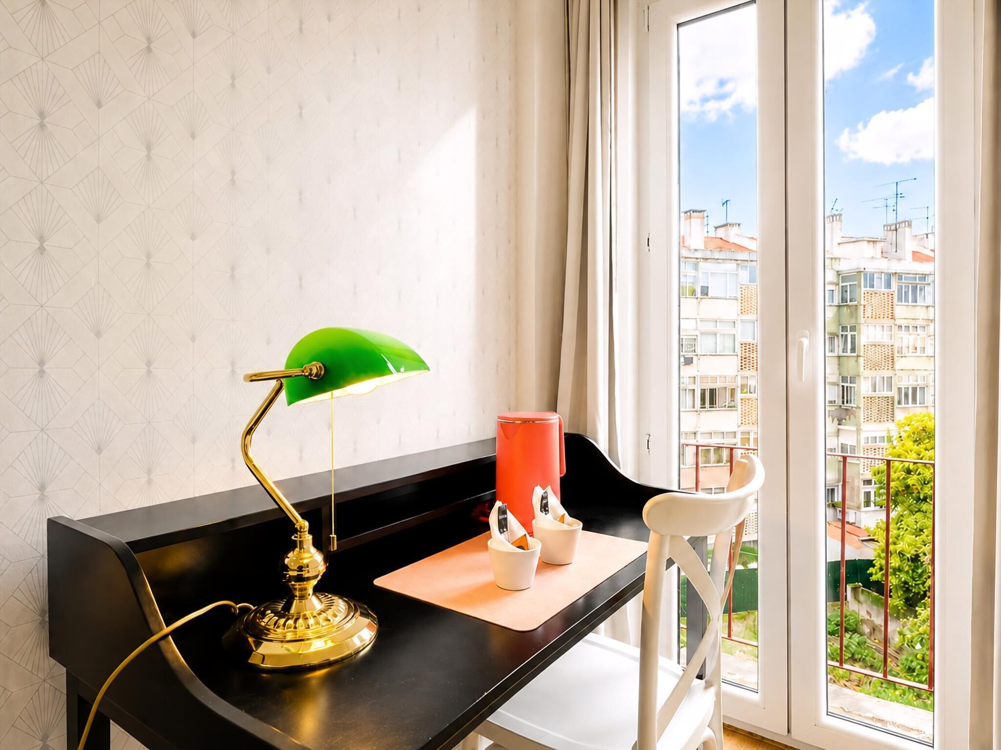 A room with a desk, a green desk lamp, a red cup, and a view of buildings through a window.