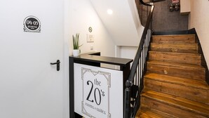 Una escalera con escalones de madera y barandillas negras, una puerta blanca con un cartel que indica '04-05 Quartos Rooms', y un cartel en la pared que anuncia 'the 20's rooms & suites'.