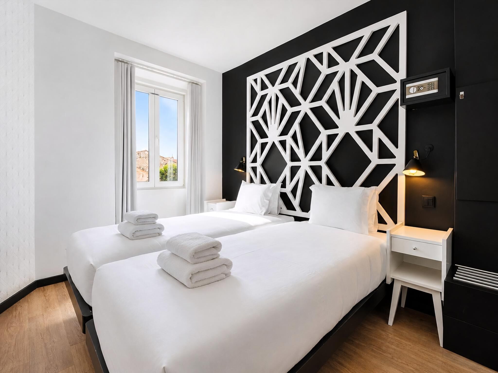Une chambre d’hôtel avec deux lits, un motif géométrique sur le mur, une fenêtre avec des rideaux et une table de chevet avec une lampe.