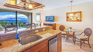 A kitchen with a large island, a dining area with wicker chairs, and a view of the ocean. - Stay Beachfront, Explore Maui’s Outdoors 629 LS (Lahaina)