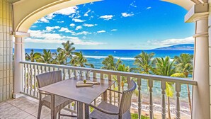 A balcony with a view of the ocean and palm trees, a table with a book and a glass, and a white railing. - Stay Beachfront, Explore Maui’s Outdoors 629 LS (Lahaina)
