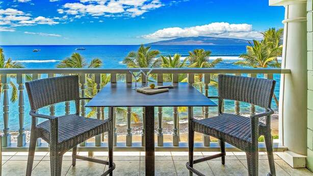 A balcony with wicker chairs and a table, overlooking a beach with palm trees and the ocean. - Stay Beachfront, Explore Maui’s Outdoors 629 LS (Lahaina)