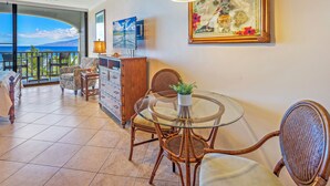 A spacious living area with a glass table, wicker chairs, a wooden cabinet, and a large painting. - Stay Beachfront, Explore Maui’s Outdoors 629 LS (Lahaina)