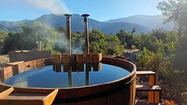 A hot tub with a wooden structure and steam rising, surrounded by a natural landscape with mountains in the background. - Espacio Arenales (Paine)