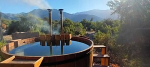 A hot tub with a wooden structure and steam rising, surrounded by a natural landscape with mountains in the background. - Espacio Arenales (Paine)