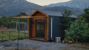 A small, modern cabin with a wooden front door and a metal roof, surrounded by mountains. - Espacio Arenales (Paine)