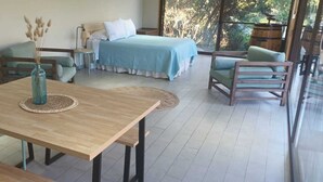 A room with a wooden table, a bed with a blue bedspread, and a view of trees through large windows. - Espacio Arenales (Paine)