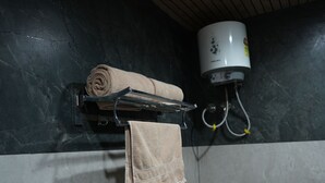 A bathroom with a towel rack holding a rolled towel and a hanging towel, a water heater, and a tiled wall. - The samsara Hotel by Satyug (Ujjain)