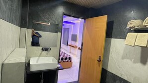A compact bathroom with a toilet, sink, and mirror. A wooden door leads to a bedroom with a bed and a TV. - The samsara Hotel by Satyug (Ujjain)