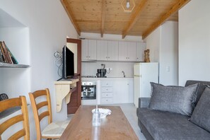 A compact living space with a wooden dining table, two wooden chairs, a grey sofa, a kitchenette with white cabinets, and a wooden ceiling.