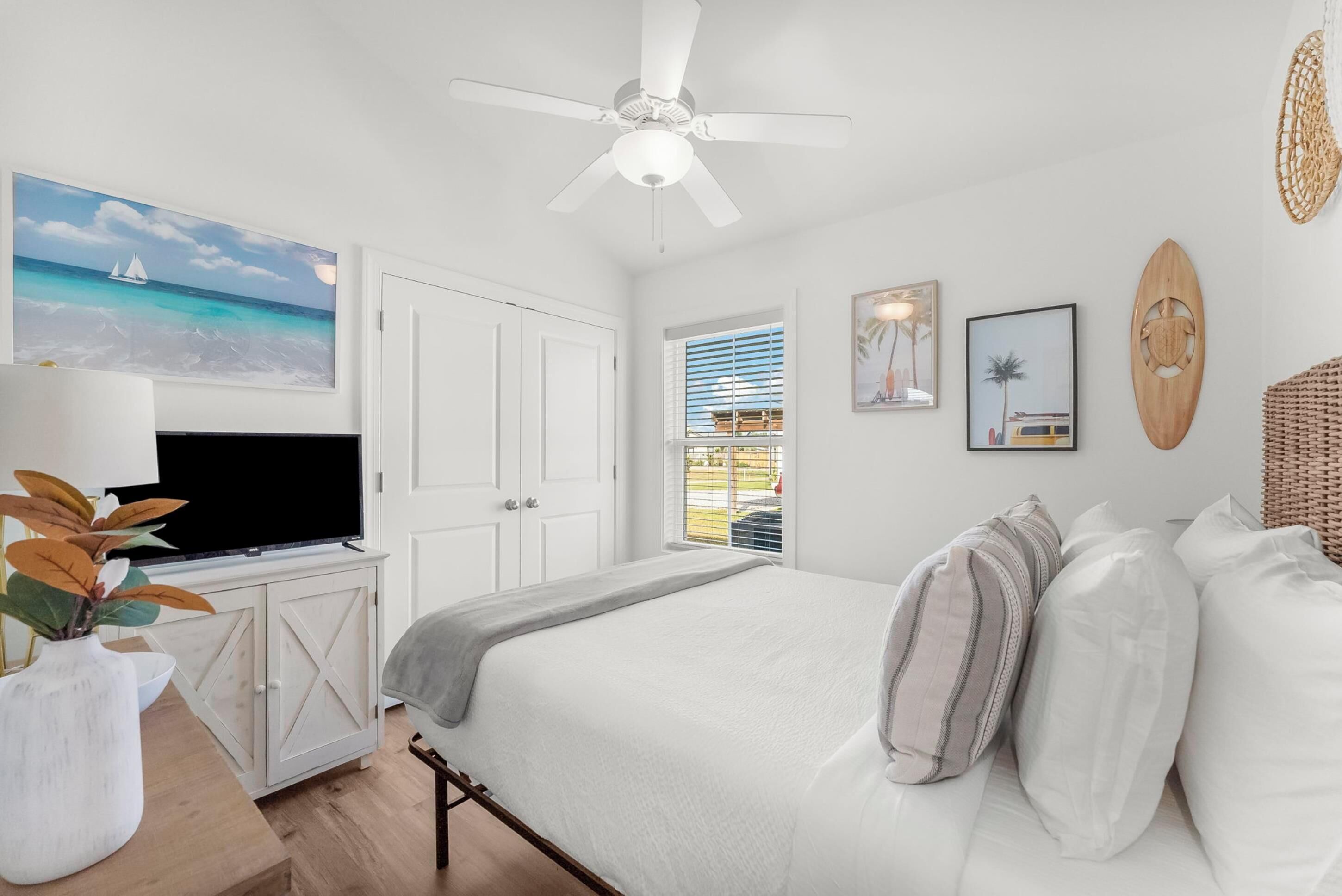 Een slaapkamer met een bed, een plafondventilator, een televisie op een kast, een raam met zonwering en wanddecoratie.