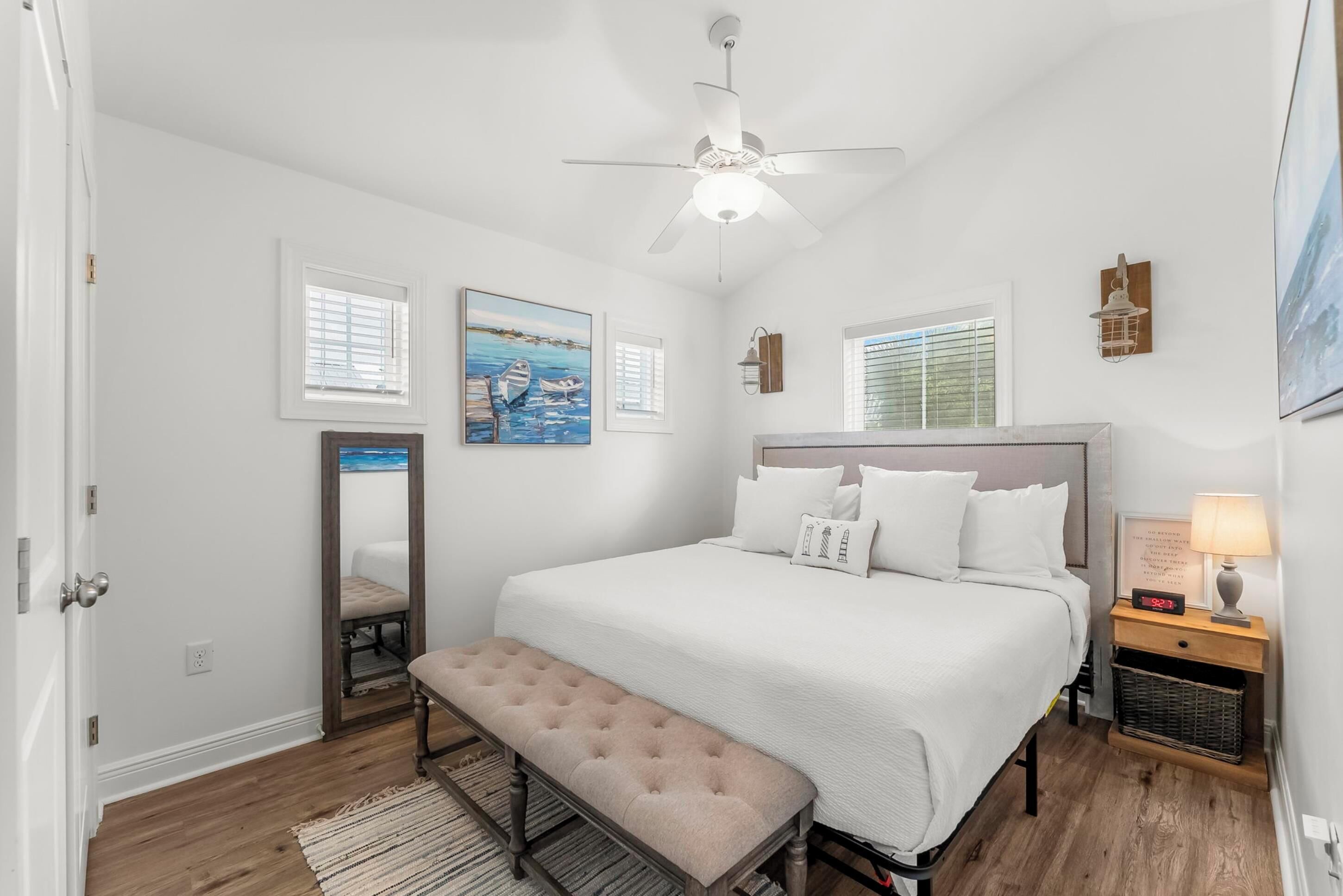 A bedroom with a large bed, a bench, a ceiling fan, a nightstand with a lamp, and artwork on the walls.