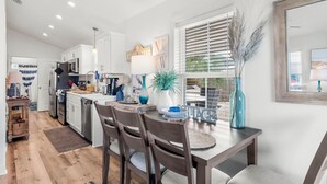 A modern kitchen with white cabinets, a dining table with chairs, and a large window with blinds. - Beach House - Sun-kissed Cottage by PHG (Port St. Joe)