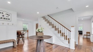 Une pièce spacieuse avec un plancher en bois, une table ronde ornée d’un vase, un escalier avec des rampes blanches et un coin repas avec des chaises.