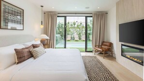 Habitación de hotel moderna con una cama grande, un televisor de pantalla plana, un área de descanso con una silla y vistas a un jardín y piscina a través de una puerta corrediza de vidrio.