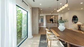 Un comedor moderno con una larga mesa de madera, sillas tejidas y un ventanal con cortinas traslúcidas.