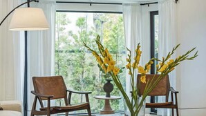 Una sala moderna con un sofá, dos sillas de madera, una mesa de centro con libros y un jarrón de flores, y un ventanal con vistas a zonas verdes.