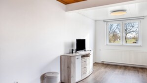 Una habitación con un armario de madera, un taburete gris, un televisor de pantalla plana y un ventanal con vistas a árboles.