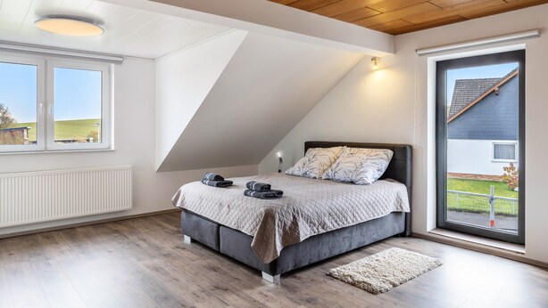 Un dormitorio con techo inclinado, cama con cabecera, radiador, ventana y vista a una casa.