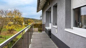 Un edificio moderno con balcón, una ventana con persianas y vistas a árboles y colinas.