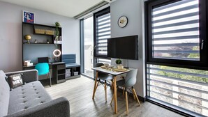 Ein modernes Wohnzimmer mit einer Couch, einem Esstisch mit Stühlen, einem Fernseher und einem großen Fenster mit Jalousien.