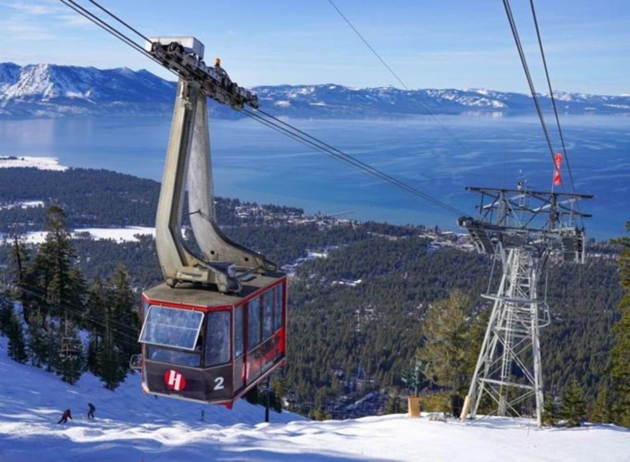A cable car with a cabin marked "H 2" suspended by cables, overlooking a snowy landscape with a lake and mountains in the distance.