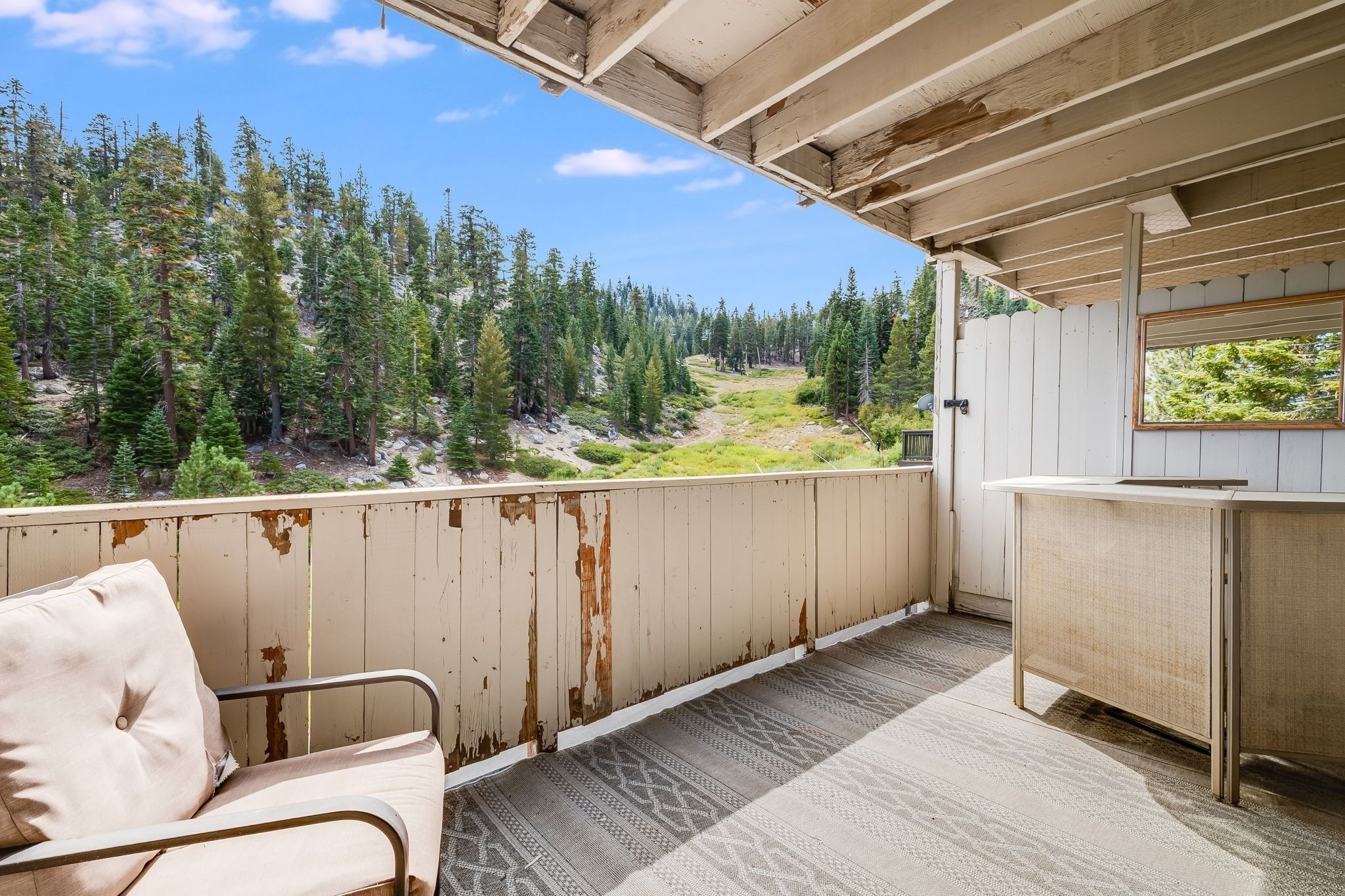A balcony with a view of a forested area, a wooden table, and a metal chair.
