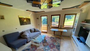 A living room with a sofa, coffee table, rug, TV, and windows with a view of greenery. - Apartment Sonnenblick with Mountain Views, Shared Garden and Wi-Fi (Bad Rippoldsau-Schapbach)