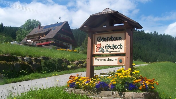A sign for a guesthouse with a wooden structure and a flower bed in front. - Apartment Sonnenblick with Mountain Views, Shared Garden and Wi-Fi (Bad Rippoldsau-Schapbach)