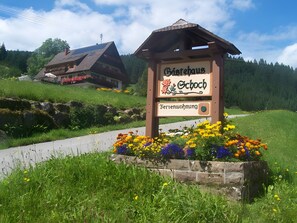 Schild für ein Gästehaus mit Holzbalkon und Blumenbeet davor.