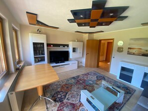 Ein modernes Wohnzimmer mit Flachbildfernseher, einem Holzessttisch, einem gemusterten Teppich und einer weißen Unterhaltungsanlage.