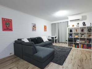 Ein Wohnzimmer mit einem grauen Ecksofa, einem kleinen weißen Tisch, grauen Vorhängen und einem Bücherregal voller Bücher und Figuren.