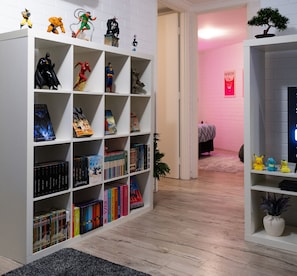 Ein Zimmer mit einem Bücherregal voller Bücher und Figuren, einer kleinen Pflanze und einem Bett im Hintergrund.