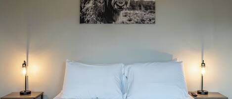 A bedroom with a bed, white pillows, two bedside tables with lamps, and a wall-mounted picture of a yak.