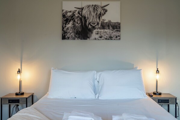 A neatly made bed with white linens and pillows, flanked by two bedside tables with lamps, in a room with a simple, clean design.