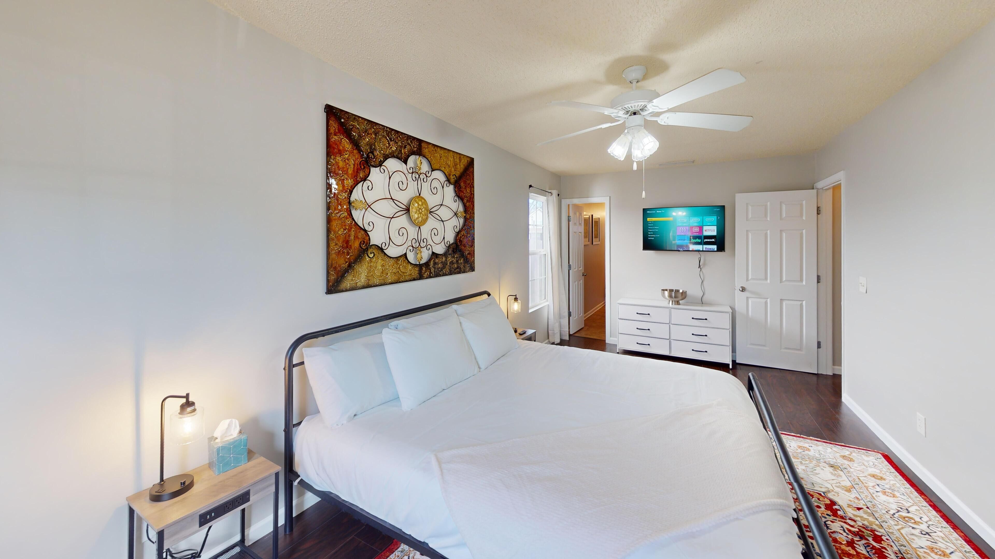 A bedroom with a bed, bedside table, lamp, artwork, ceiling fan, TV, dresser, and a door leading to another room.