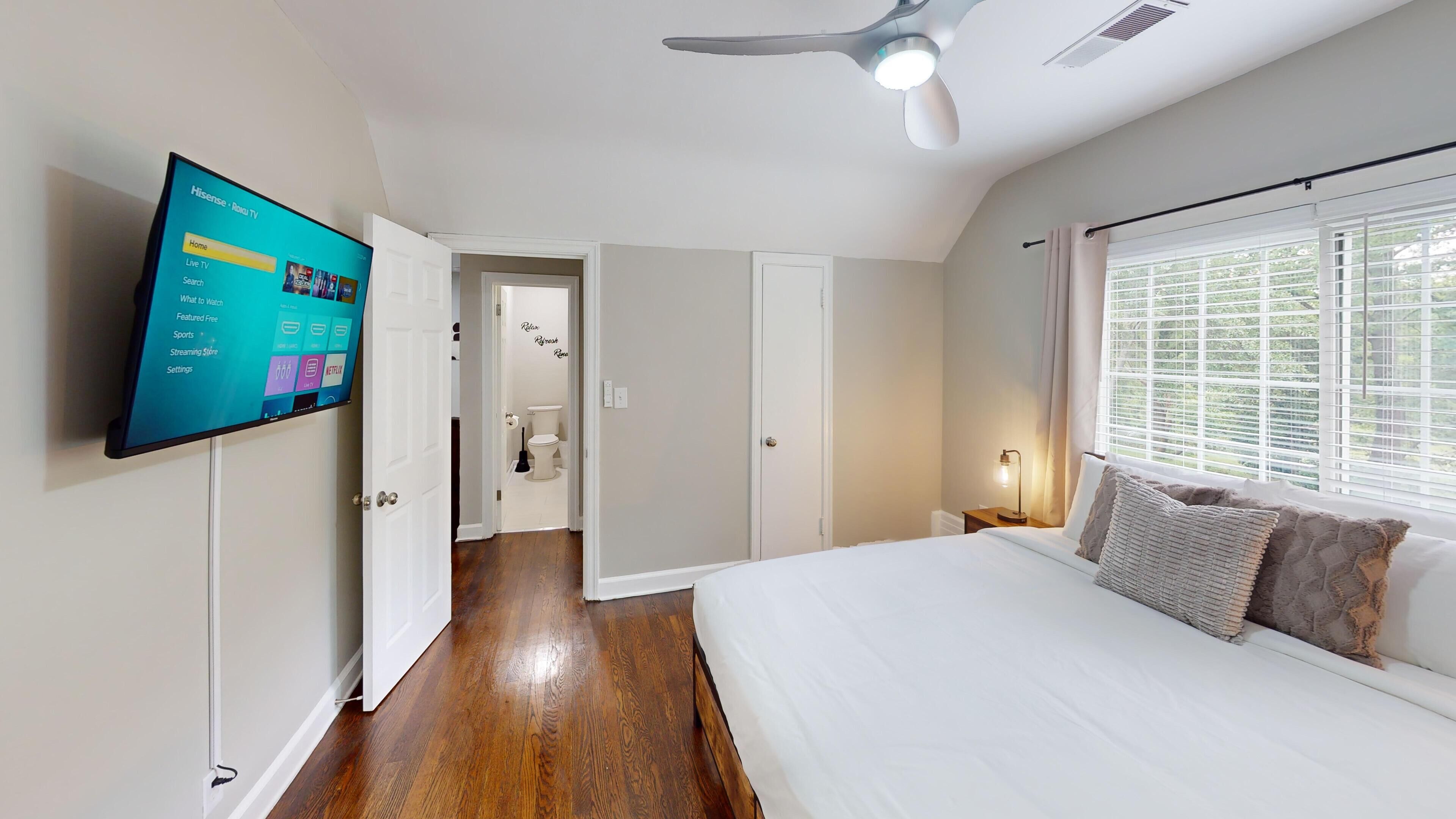 Une chambre à coucher avec un lit, un téléviseur fixé au mur, un ventilateur de plafond et une fenêtre avec des stores.