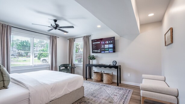 A modern bedroom with a bed, a ceiling fan, a TV on the wall, and a view of the outside through large windows. - 18 Huntcliff Dr, Cartersville (Cartersville)