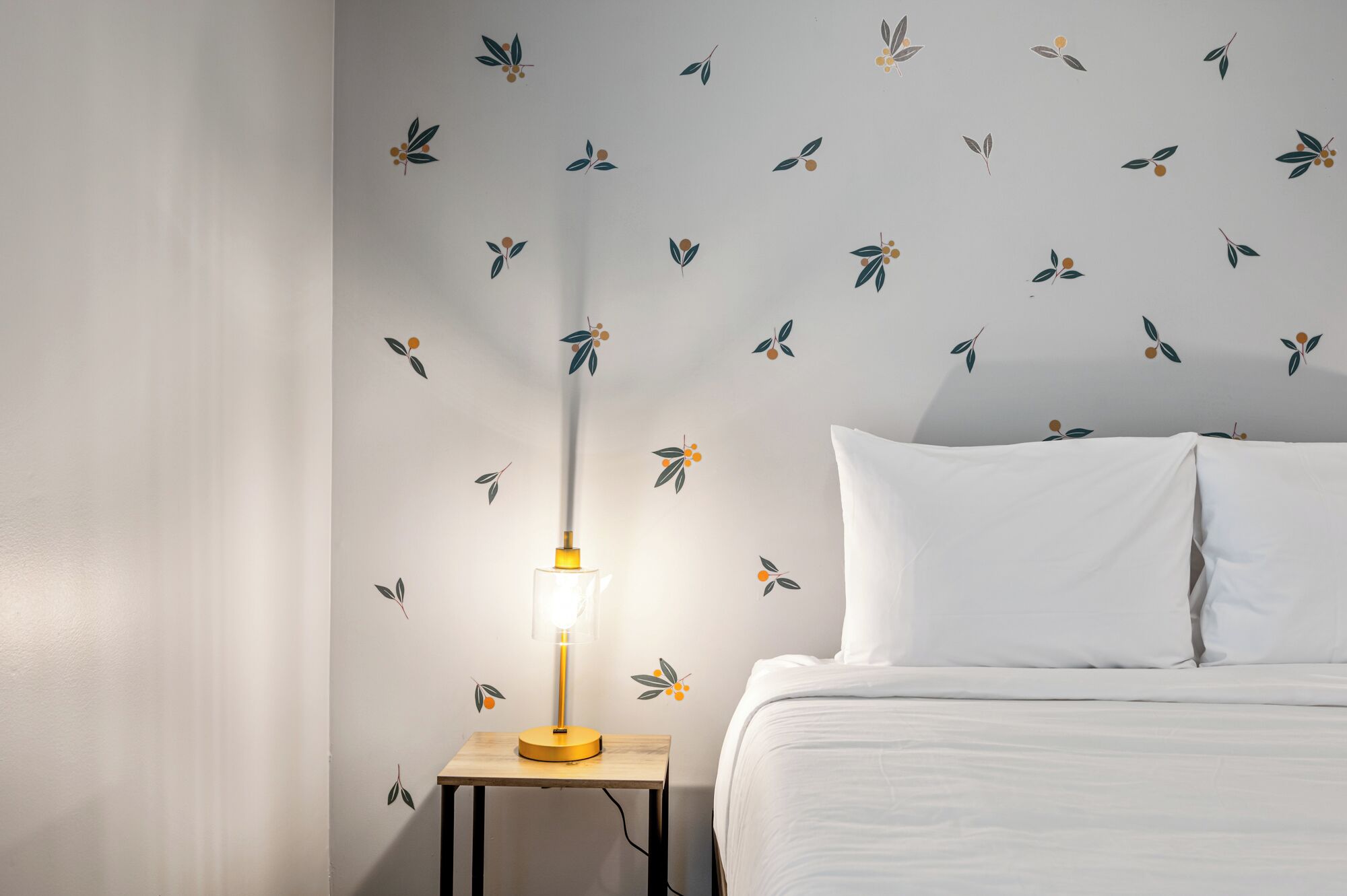 Een slaapkamer met een bed, witte kussens, een nachtkastje met een lamp en een muur met een bladmotief.