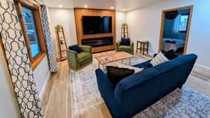 A modern living room with a flat-screen TV, green and blue sofas, a wooden coffee table, and a patterned rug.