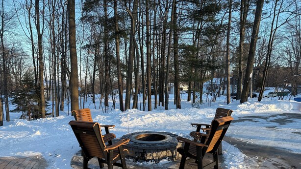 A wooden deck with two Adirondack chairs and a fire pit in the center, surrounded by snow and trees.