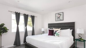 Ein modernes Schlafzimmer mit einem großen Bett, grauen Vorhängen, einer Topfpflanze und einem gerahmten Kunstwerk an der Wand.