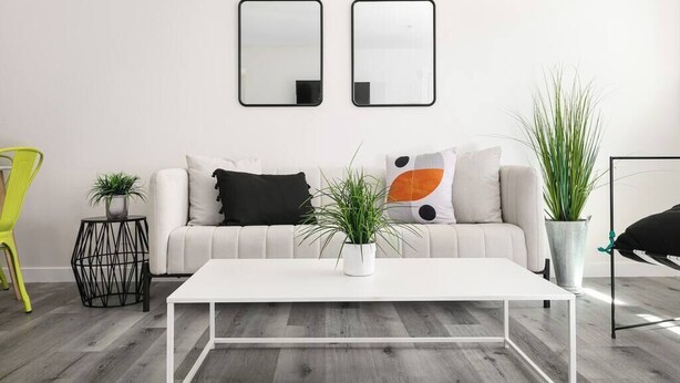Ein modernes Wohnzimmer mit einem weißen Sofa, zwei Spiegeln mit schwarzem Rahmen, einem weißen Couchtisch und Topfpflanzen.
