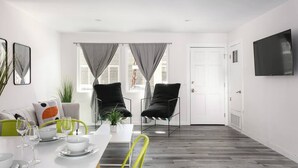 Ein modernes Wohnzimmer mit Essbereich, ausgestattet mit einem weißen Sofa, zwei schwarzen Sesseln, einem Esstisch mit Gläsern und einem flachbildfernseher an der Wand.